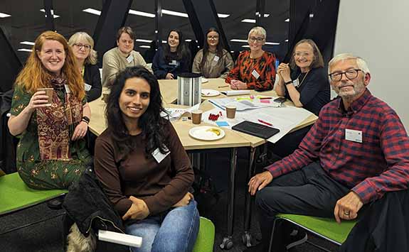 Nine members of the One Community Network sat around a table at an event. All casually dressed and smiling at the camera. 